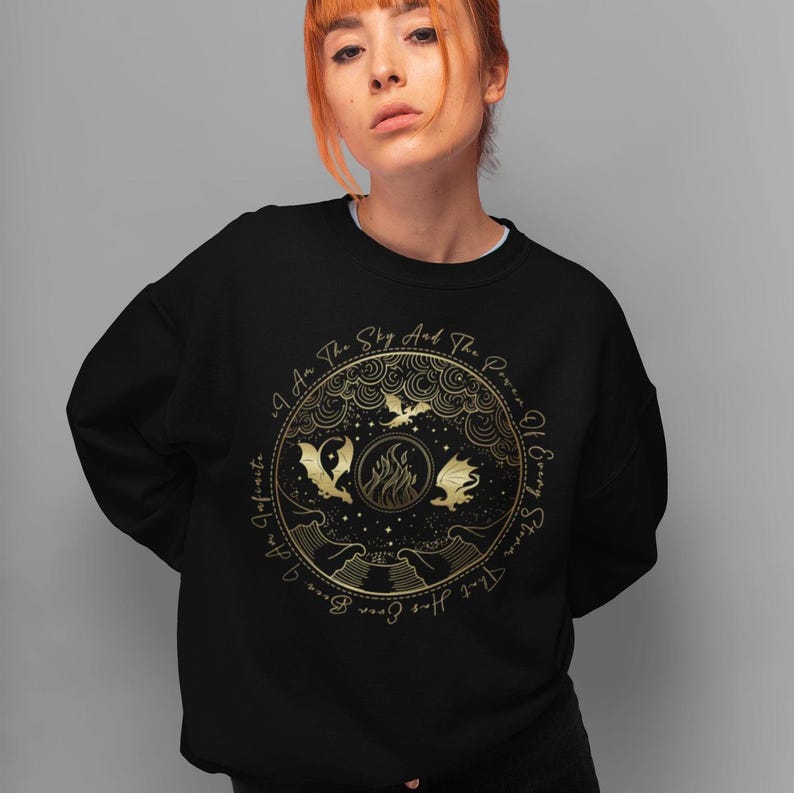 K&ouml;nnte beinhalten: Schwarzer Pullover mit einem goldenen Design aus drei Drachen, die um ein Feuer fliegen. Der Text um das Design lautet "Ich bin der Himmel und das Feuer des Alten und des Neuen."