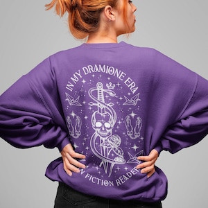 K&ouml;nnte beinhalten: Lilafarbener Pullover mit einem wei&szlig;en Grafikdruck eines Sch&auml;dels, einer Schlange und eines Schwertes. Der Text "In My Dramione Era" und "In Fiction Reader" ist auf dem Pullover aufgedruckt.