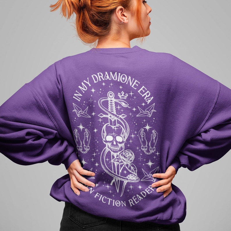 K&ouml;nnte beinhalten: Lilafarbener Pullover mit einem wei&szlig;en Grafikdruck eines Sch&auml;dels, einer Schlange und eines Schwertes. Der Text "In My Dramione Era" und "In Fiction Reader" ist auf dem Pullover aufgedruckt.