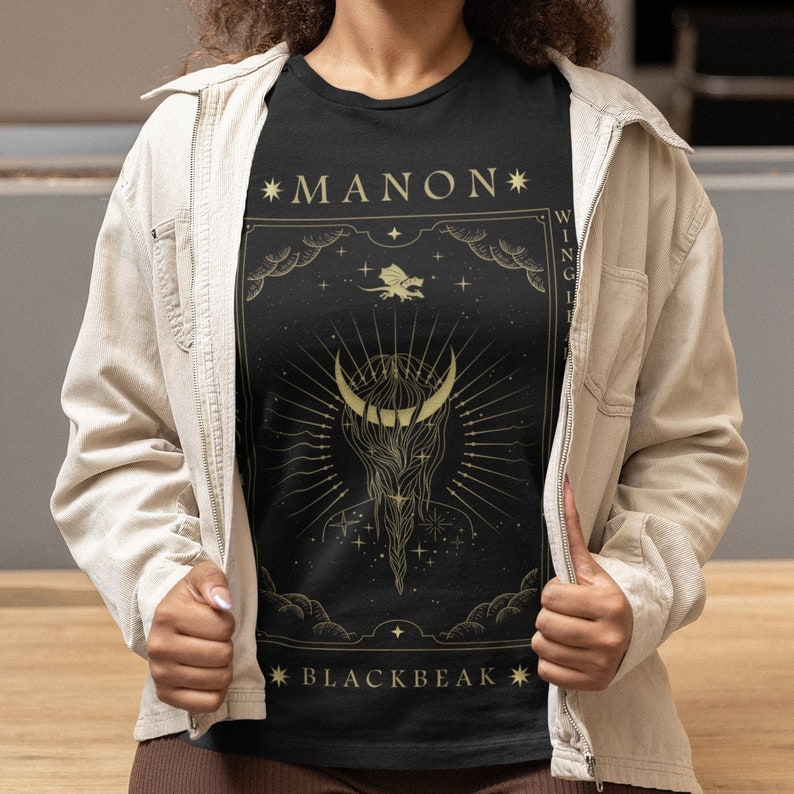 May include: Black t-shirt with a gold graphic design featuring a crescent moon, stars, and the text "*MANON*" and "*BLACKBEAK*"