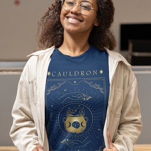 May include: A navy blue t-shirt with a gold graphic of a cauldron with a crescent moon and stars. The text "*CAULDRON*" and "*BLESSED*" is printed in gold on the shirt.