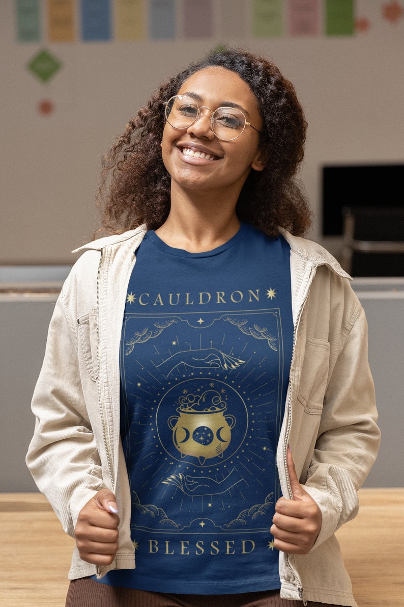 May include: A navy blue t-shirt with a gold graphic of a cauldron with a crescent moon and stars. The text "*CAULDRON*" and "*BLESSED*" is printed in gold on the shirt.