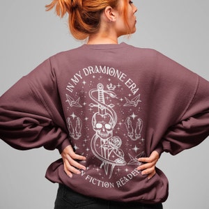K&ouml;nnte beinhalten: Ein maroonfarbener Pullover mit einem wei&szlig;en Grafikdruck eines Sch&auml;dels, einer Schlange und eines Schwertes. Der Text auf der Grafik lautet "In My Dramione Era" und "In Fiction Reader".