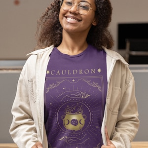 May include: Purple t-shirt with a gold graphic of a cauldron with stars and moons. The text "*CAULDRON*" and "*BLESSED*" is printed on the shirt.
