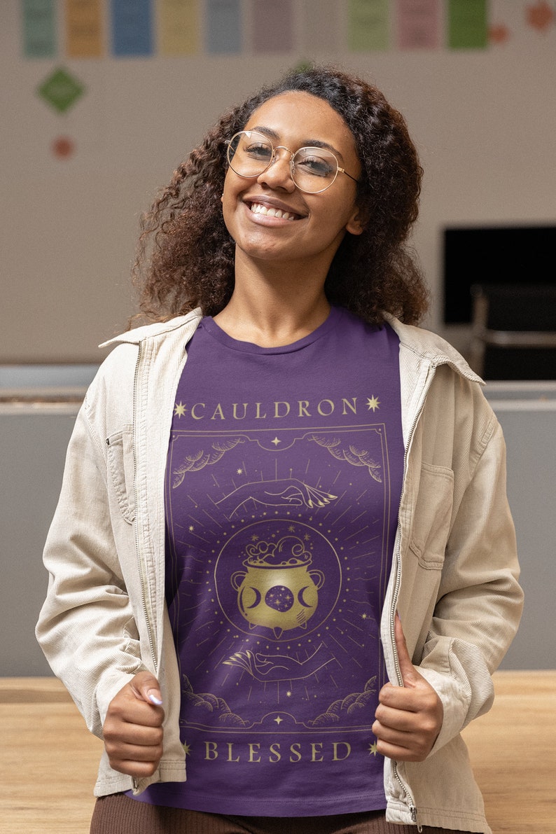 May include: Purple t-shirt with a gold graphic of a cauldron with stars and moons. The text "*CAULDRON*" and "*BLESSED*" is printed on the shirt.