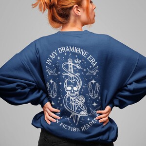 K&ouml;nnte beinhalten: Ein marineblauer Pullover mit einem wei&szlig;en Grafikdesign, das einen Sch&auml;del, eine Schlange und ein Schwert zeigt. Der Text auf der Grafik lautet "In My Dramione Era" und "An Fiction Reader".