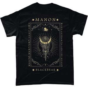 May include: Black t-shirt with a gold graphic design featuring a crescent moon, stars, and the text "*MANON* THE LAST CROCHAN QUEEN WING LEADER OF THE THIRTEEN *BLACKBEAK*"