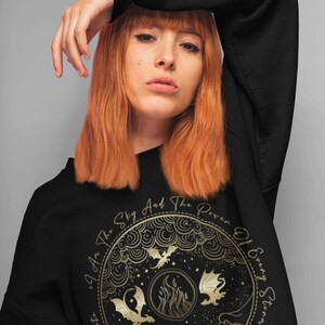 K&ouml;nnte beinhalten: Schwarzes Sweatshirt mit einem goldenen Grafikdruck eines Kreises mit Drachen, Flammen und Sternen. Der Text "I Am The Sky And The Power Of Every Storm That Has Ever Been" ist um den Kreis herum gedruckt.