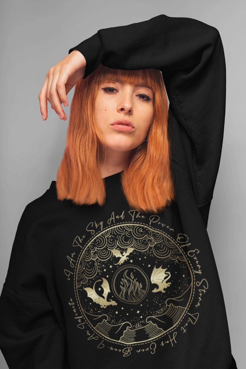K&ouml;nnte beinhalten: Schwarzes Sweatshirt mit einem goldenen Grafikdruck eines Kreises mit Drachen, Flammen und Sternen. Der Text "I Am The Sky And The Power Of Every Storm That Has Ever Been" ist um den Kreis herum gedruckt.