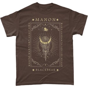May include: A brown t-shirt with a gold graphic design featuring a crescent moon, stars, and the text "*MANON* THE LAST CROCHAN QUEEN WING LEADER OF THE THIRTEEN *BLACKBEAK*"