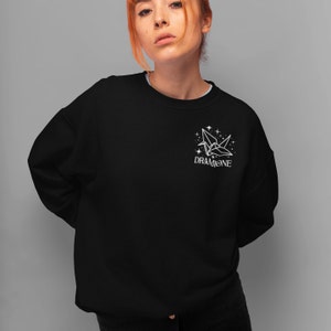 K&ouml;nnte beinhalten: Schwarzer Pullover mit einem wei&szlig;en Origami-Kranich und Sternen-Motiv auf der linken Brust. Der Text "DRAMIONE" ist unter dem Motiv gedruckt.