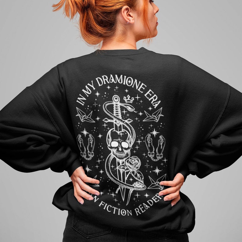 K&ouml;nnte beinhalten: Schwarzer Pullover mit einem wei&szlig;en Grafikdruck eines Sch&auml;dels mit einer Schlange, die um einen Dolch gewickelt ist. Der Text "In My Dramione Era" und "A Fiction Reader" ist um die Grafik gedruckt.