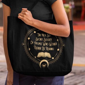 Op de afbeelding: Zwarte draagtas met een goud en wit ontwerp. Het ontwerp toont de tekst "The Not So Secret Society Of People Who Would Rather Be Reading" rond een open boek en een halve maan. De tas wordt door een persoon vastgehouden.