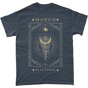 May include: A dark blue t-shirt with a gold graphic design featuring a crescent moon, stars, and the text "*MANON* THE LAST CROCHAN QUEEN WING LEADER OF THE THIRTEEN *BLACKBEAK*"