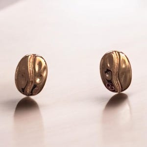 May include: Two gold-colored, oval-shaped earrings. Each earring has a vertical, darker stripe down the center and a reflective surface. The earrings are displayed against a light background.
