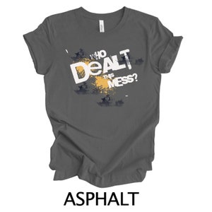May include: A dark gray t-shirt with a white graphic that says "Who Dealt This Mess?" The graphic is surrounded by a yellow and black splatter design.