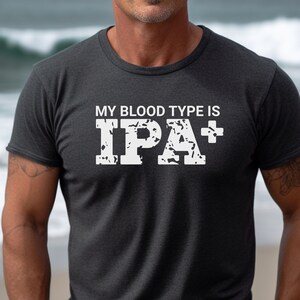 May include: Dark grey t-shirt featuring the white text "MY BLOOD TYPE IS IPA+" in a distressed font. The shirt has a crew neck and short sleeves. The background is a blurred view of the sea.