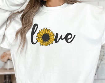 Op de afbeelding: Witte trui met een zwarte en gele zonnebloem afbeelding en het woord "love" gedrukt op de voorkant.