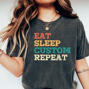 May include: A dark grey t-shirt with a vintage-style graphic that reads "Eat Sleep Custom Repeat" in rainbow colours.