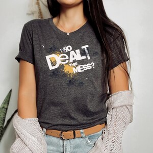 May include: A dark gray t-shirt with a white graphic that says "Who dealt this mess?" The graphic is in a yellow and white paint splatter design.