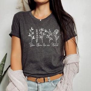 May include: A dark gray t-shirt with a white graphic of stylized flowers and the text "Bloom Where You Are Planted".