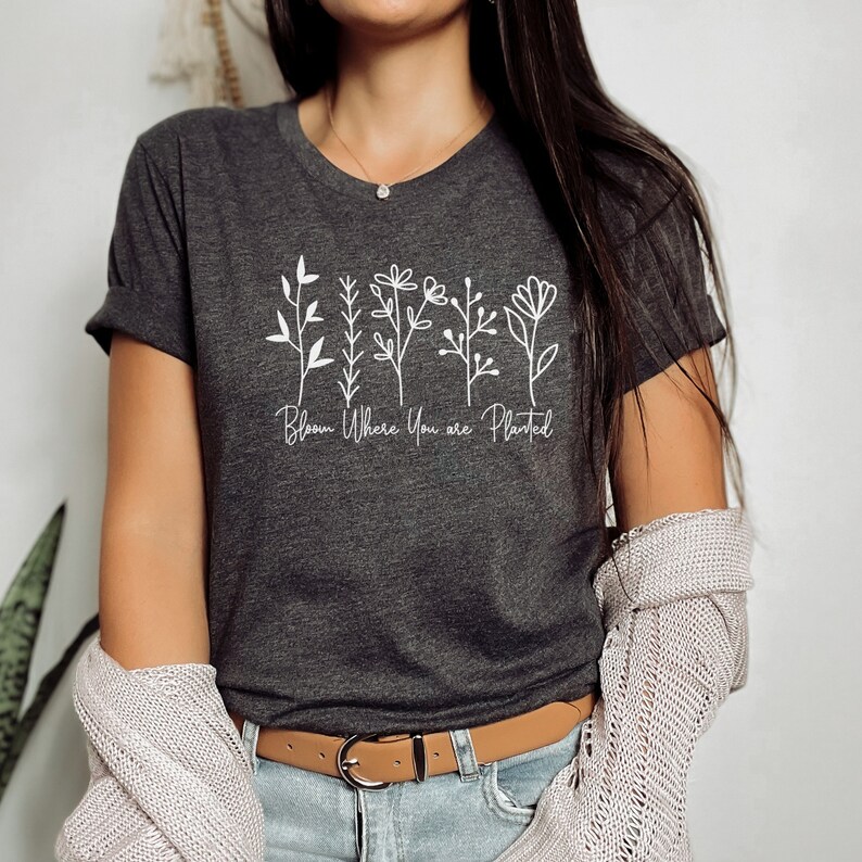 May include: A dark gray t-shirt with a white graphic of stylized flowers and the text "Bloom Where You Are Planted".