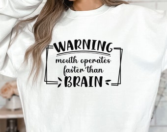 Op de afbeelding: Wit sweatshirt met zwarte tekst die "WARNING mouth operates faster than BRAIN" luidt.