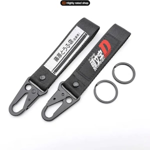 May include: Two black keychains with metal clasps and key rings. One keychain has Japanese text on a white label, the other has a red and white graphic with Japanese text. The keychains are made of a durable black fabric.