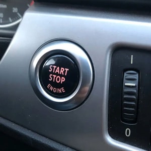 May include: Close-up of a car's dashboard featuring a black start/stop engine button with the words "START STOP ENGINE" in pink. The button is surrounded by a silver metallic trim. A black dial with white markings is visible to the right.