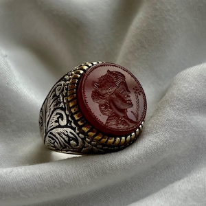 May include: A silver ring with a red cameo featuring a woman's face. The ring has a gold band and intricate floral designs.