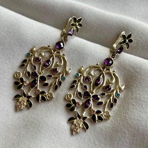 May include: A pair of gold filigree earrings featuring purple, pink and green gemstones. The earrings are shaped like a floral design.