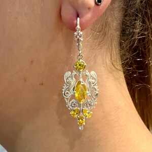 May include: Silver dangle earrings with a yellow and pink gemstone design. The earrings feature a large oval-shaped yellow gemstone surrounded by smaller yellow and pink gemstones.