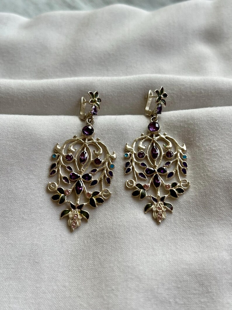 May include: A pair of silver dangle earrings with a floral design. The earrings are adorned with purple, pink, and turquoise gemstones.