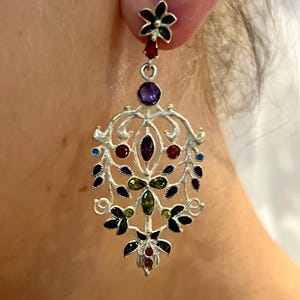 May include: Silver dangle earrings with a floral design and colorful gemstones. The earrings feature a black flower at the top, followed by a cascade of green, red, purple, and blue gemstones.