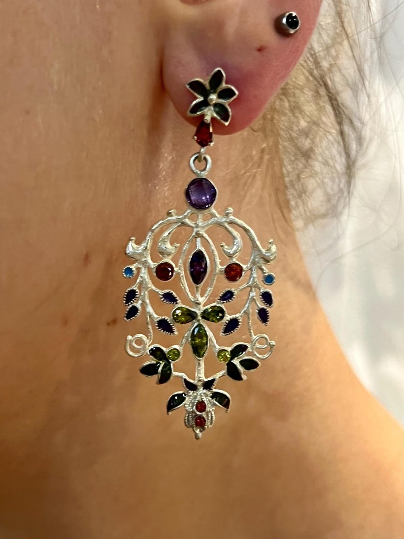 May include: Silver dangle earrings with a floral design and colorful gemstones. The earrings feature a black flower at the top, followed by a cascade of green, red, purple, and blue gemstones.