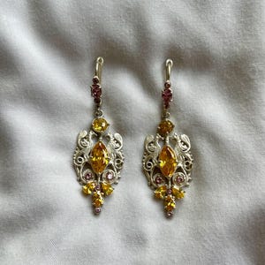 May include: A pair of silver dangle earrings with yellow and pink gemstones. The earrings feature a delicate filigree design.