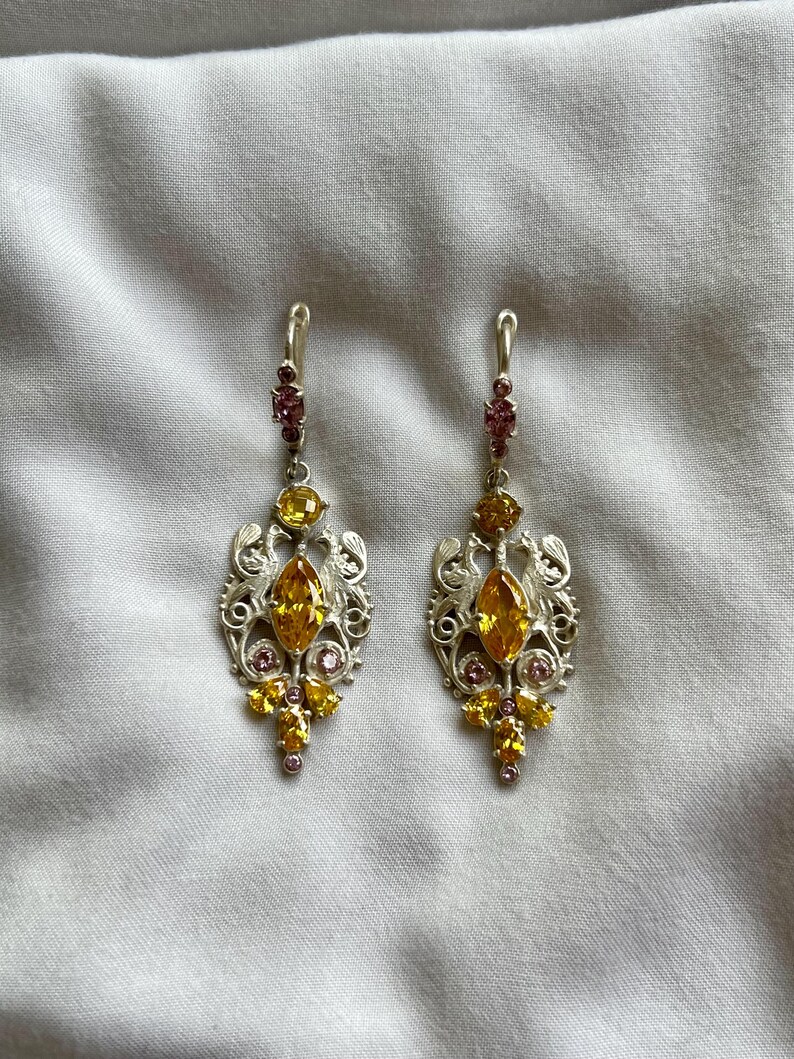 May include: A pair of silver dangle earrings with yellow and pink gemstones. The earrings feature a delicate filigree design.