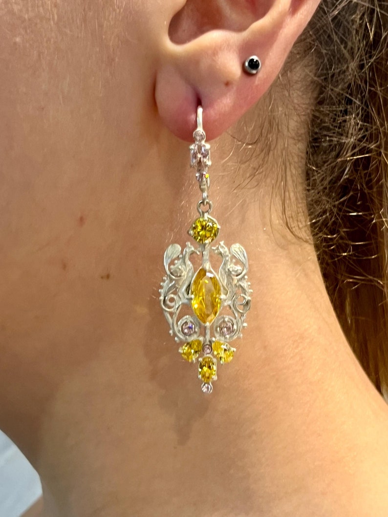 May include: Silver dangle earrings with a yellow and pink gemstone design. The earrings feature a large oval-shaped yellow gemstone surrounded by smaller yellow and pink gemstones.