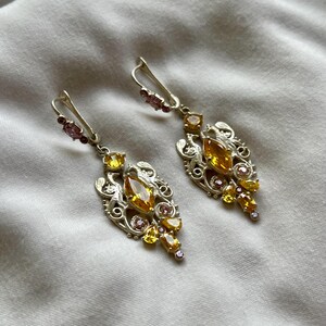 May include: A pair of silver earrings with a delicate filigree design. The earrings feature a cluster of yellow and pink gemstones.