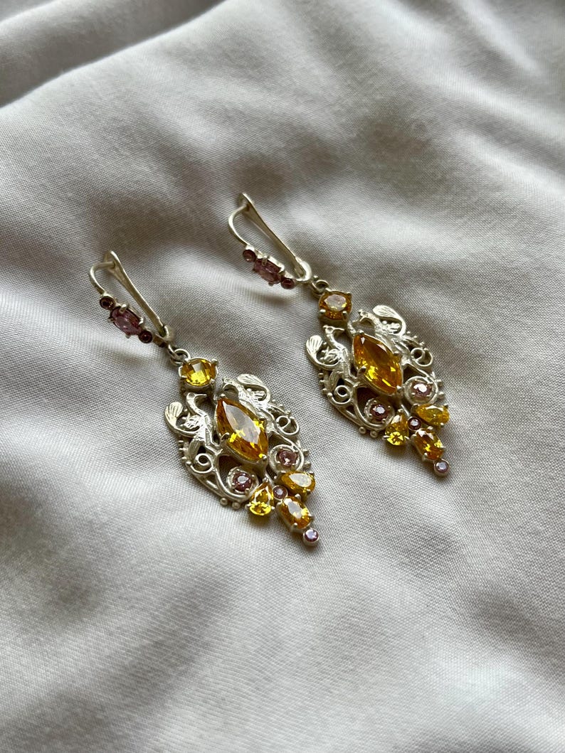 May include: A pair of silver earrings with a delicate filigree design. The earrings feature a cluster of yellow and pink gemstones.