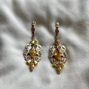 May include: A pair of silver dangle earrings with a delicate filigree design. The earrings feature multiple yellow and pink gemstones.