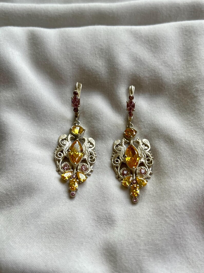 May include: A pair of silver dangle earrings with a delicate filigree design. The earrings feature multiple yellow and pink gemstones.