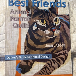 Peut inclure: Couverture de livre "Best Friends Animal Portrait Quilts" de June Jaeger. Elle présente une illustration détaillée d'un chat avec des rayures marron et noires, des yeux jaunes et des accents bleus. Le sous-titre est "Quilter's Guide to Animal Designs."