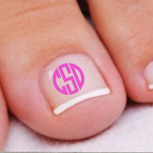 May include: Close-up of a person's toes with a pink monogram design on the big toenail. The monogram is a circular design with the letters C, S, and D. The nails are painted with a French manicure style.