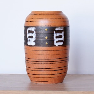 Puede incluir: Un jarrón de cerámica marrón con un patrón de rayas y diseños geométricos blancos y negros. El jarrón tiene una boca ancha y una base estrecha.
