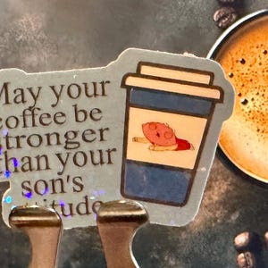 May include: A sticker with the text "May your coffee be stronger than your son's attitude" and a cartoon coffee cup illustration. The cup has a blue body and a cartoon character. A cup of coffee is in the background.