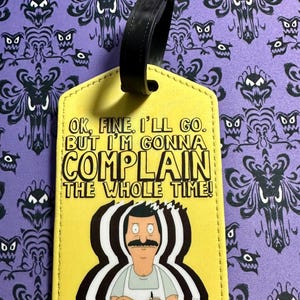 May include: A yellow rectangular luggage tag with a black strap. The tag features a graphic of Bob Belcher with his arms crossed and the text: OK, FINE. I'LL GO. BUT I'M GONNA COMPLAIN THE WHOLE TIME! The design includes a black and white motion effect.