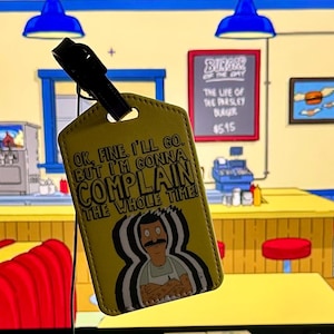 May include: Yellow luggage tag featuring a cartoon image of Bob from the animated series, with the text "OK, FINE. I'LL GO, BUT I'M GONNA COMPLAIN THE WHOLE TIME!" The tag has a black strap and is set against a cartoon restaurant background.