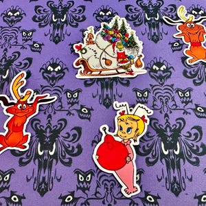 May include: A collection of colorful cartoon character stickers on a purple background with a black pattern. The stickers feature characters from a holiday story, including a sleigh, a dog-like creature, and a girl holding a red ornament.