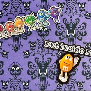 May include: A purple background with a black and white pattern featuring cartoon ghosts and ornate designs. Colorful M&M's stickers are arranged across the top, with a yellow M&M sticker below the text "nut inside me."